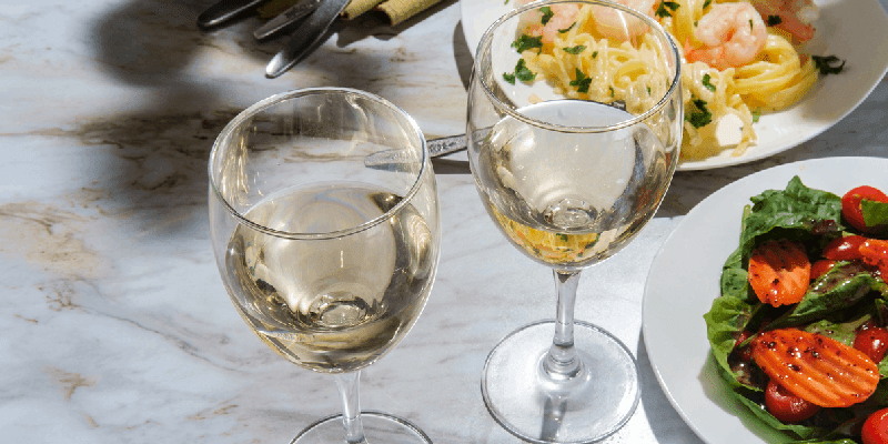 two glasses of white wine next to a plate of fettuccini alfredo